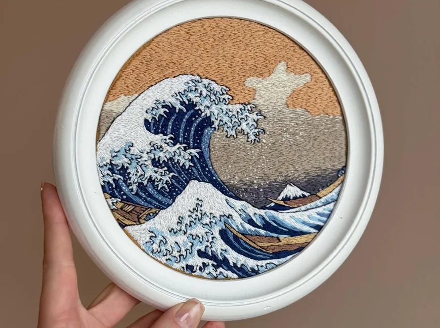 An embroidered reproduction of The Great Wave in a round white frame, held in a hand