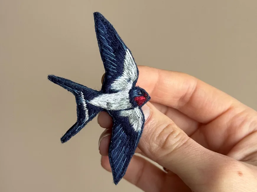 An embroidered swallow brooch in navy, white and red, held between two fingers