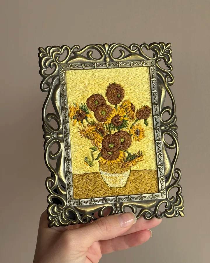 An embroidered reproduction of Van Gogh's Sunflowers in an ornate antique-gold frame, held in a hand